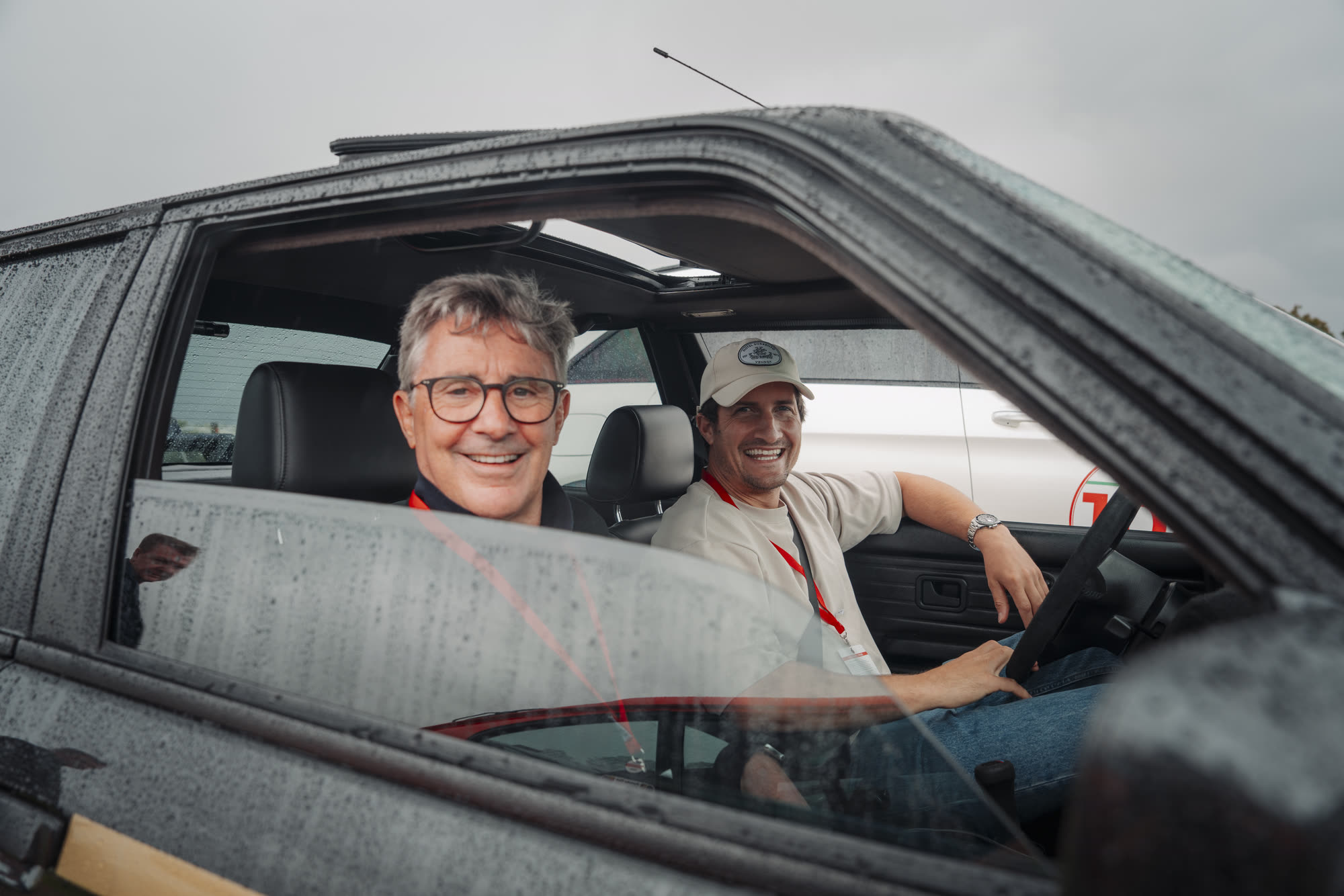 Two men grinning from the front seats of a car on a wet morning