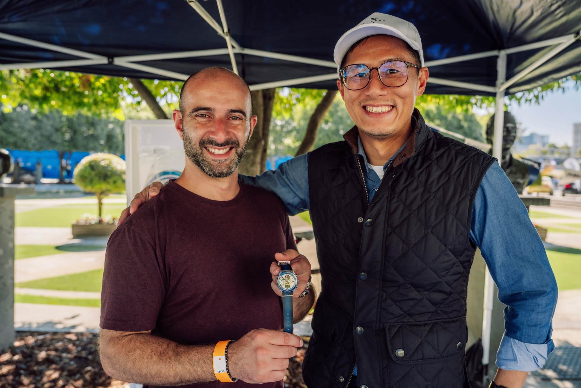 Two men under a marquee holding up a wristwatch for the camera