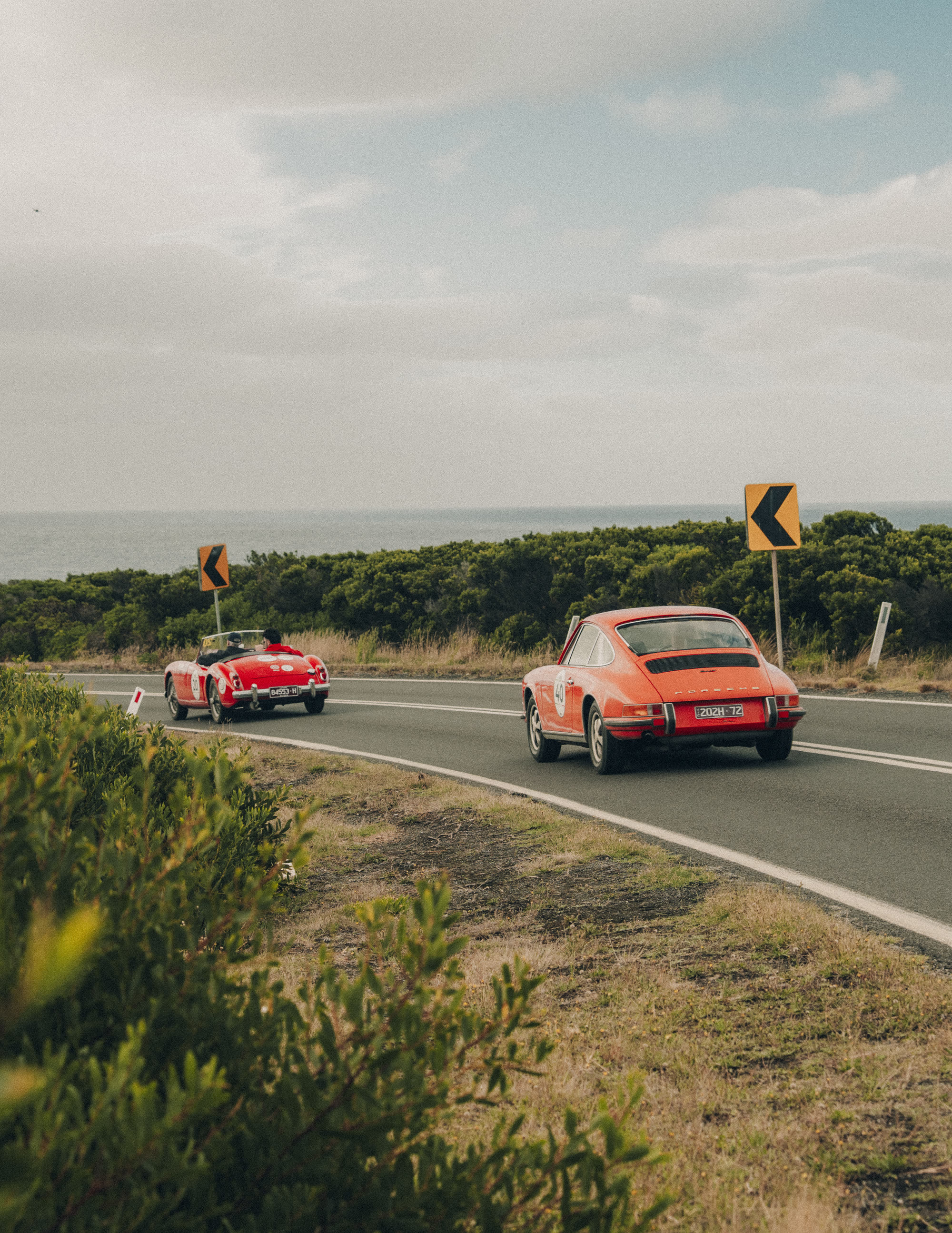 An orange Porsche 911 following a red roadster on a road above the sea