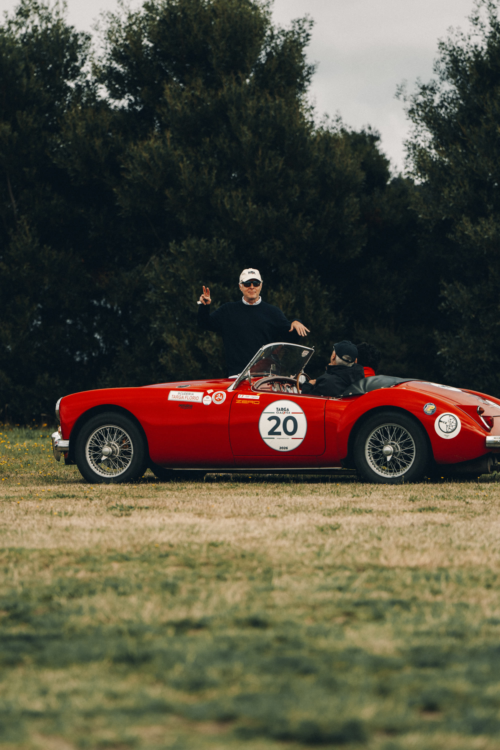 A man standing beside a red open-top classic roadster parked on grass
