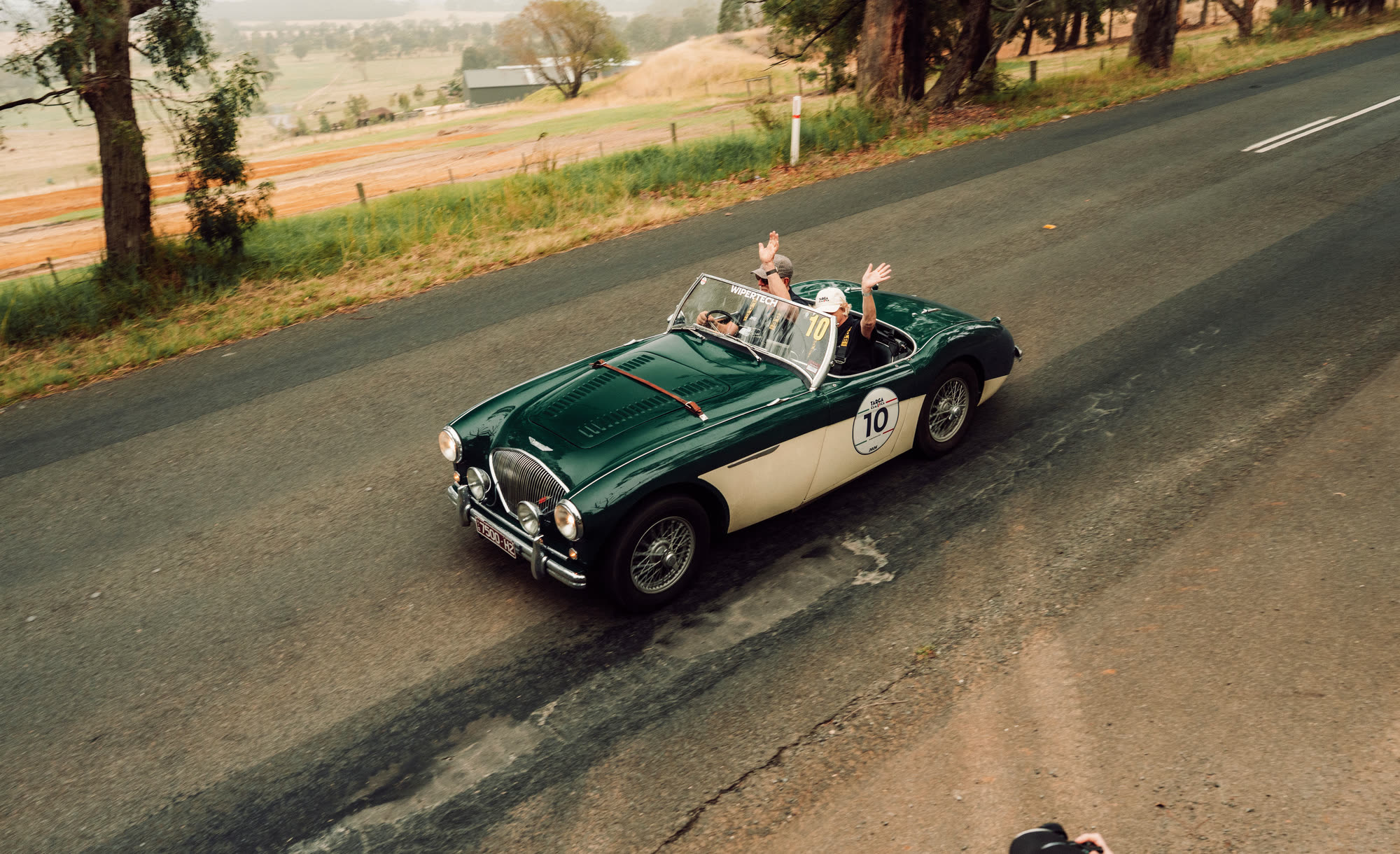 Two crew waving from a green and cream classic roadster above farmland