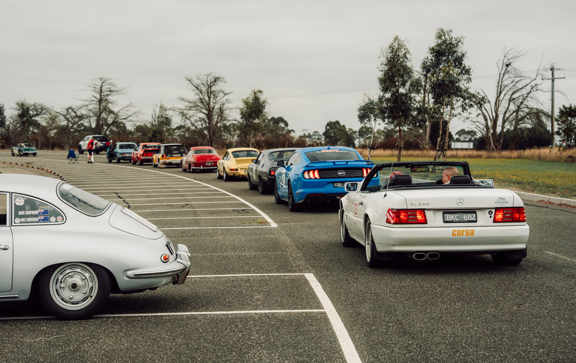 Cars queued in a rural car park behind a silver Porsche 356 coupe