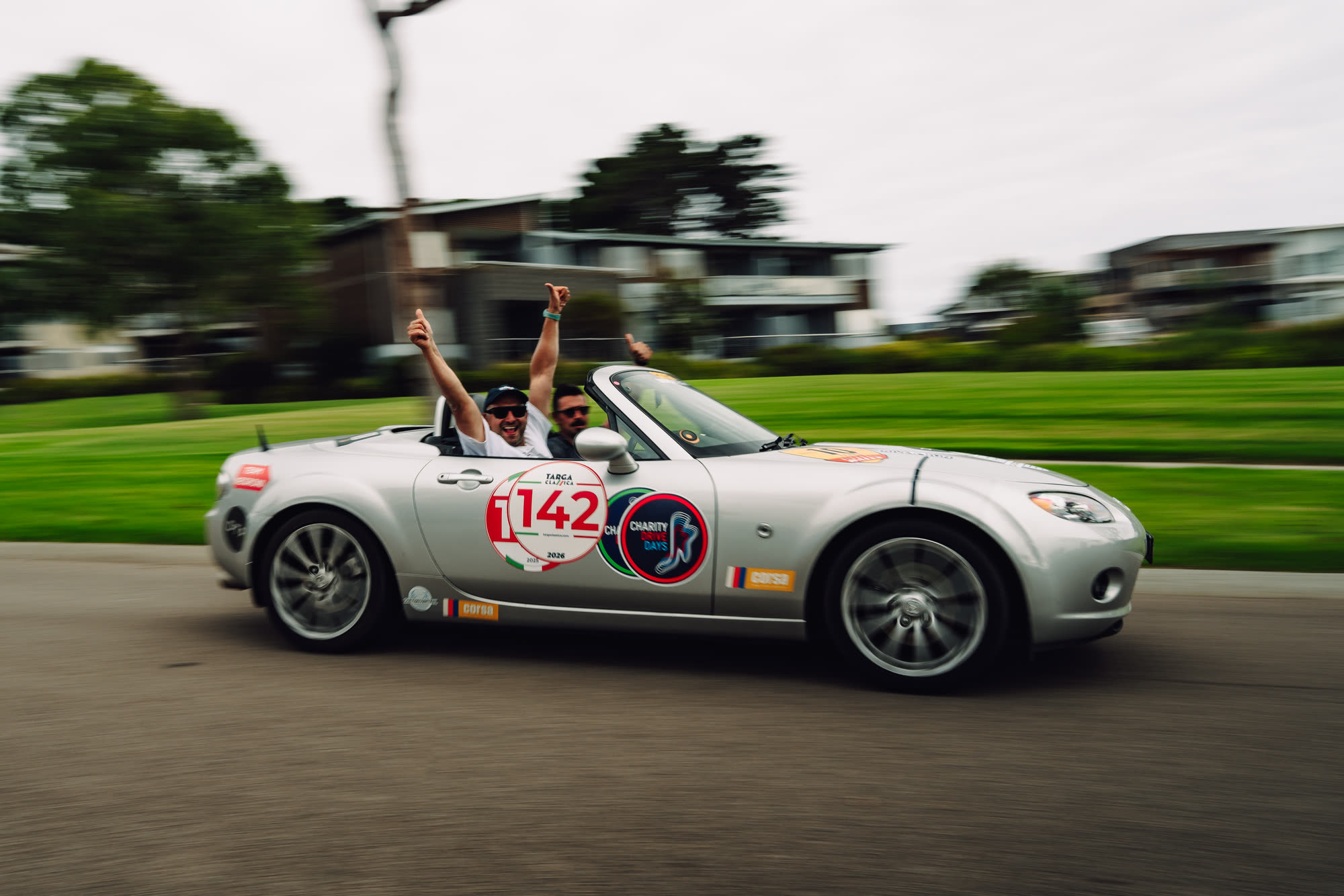 Two men cheering from a silver convertible at speed, running number 142