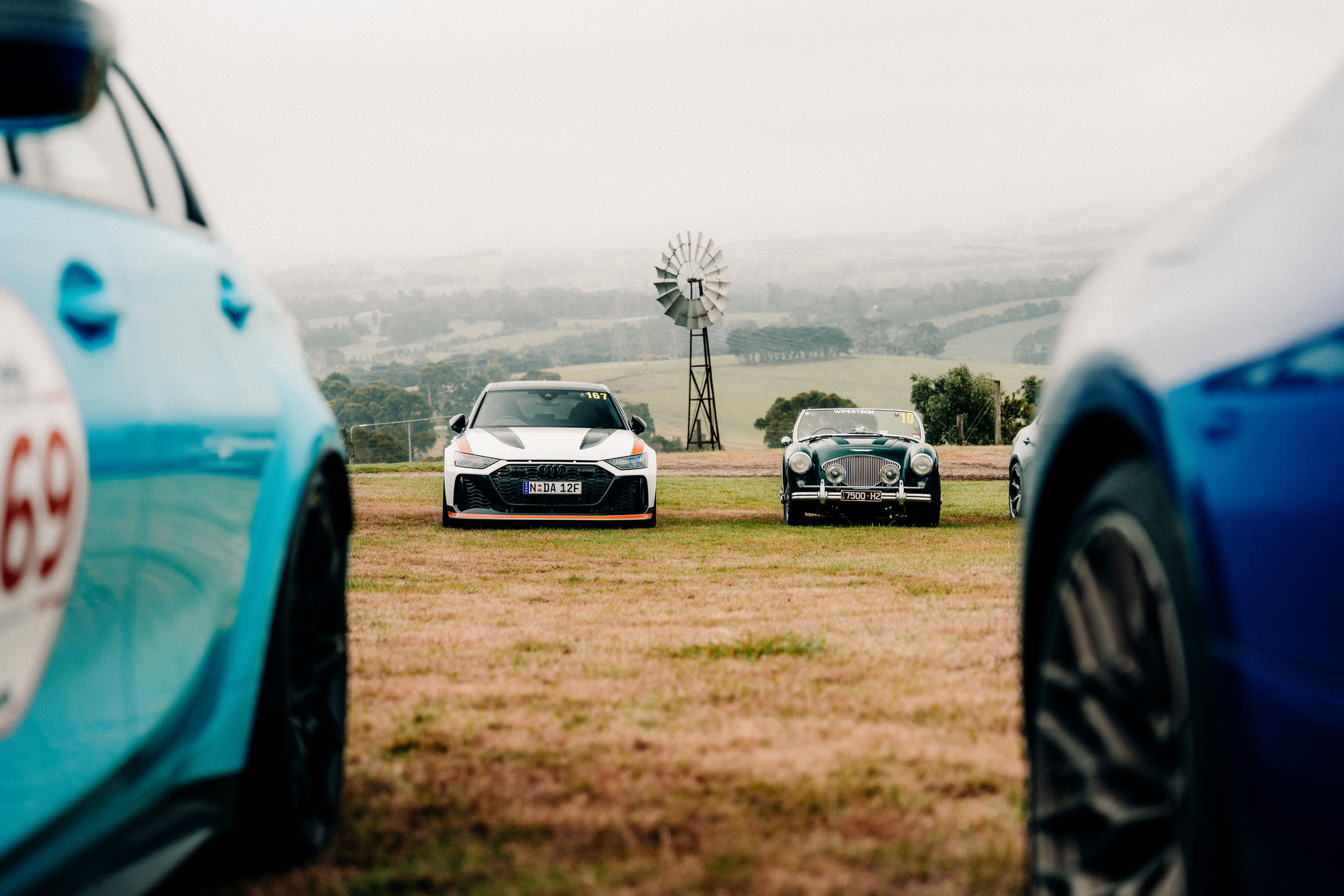 A white Audi and a green classic roadster parked on a hill, windmill behind