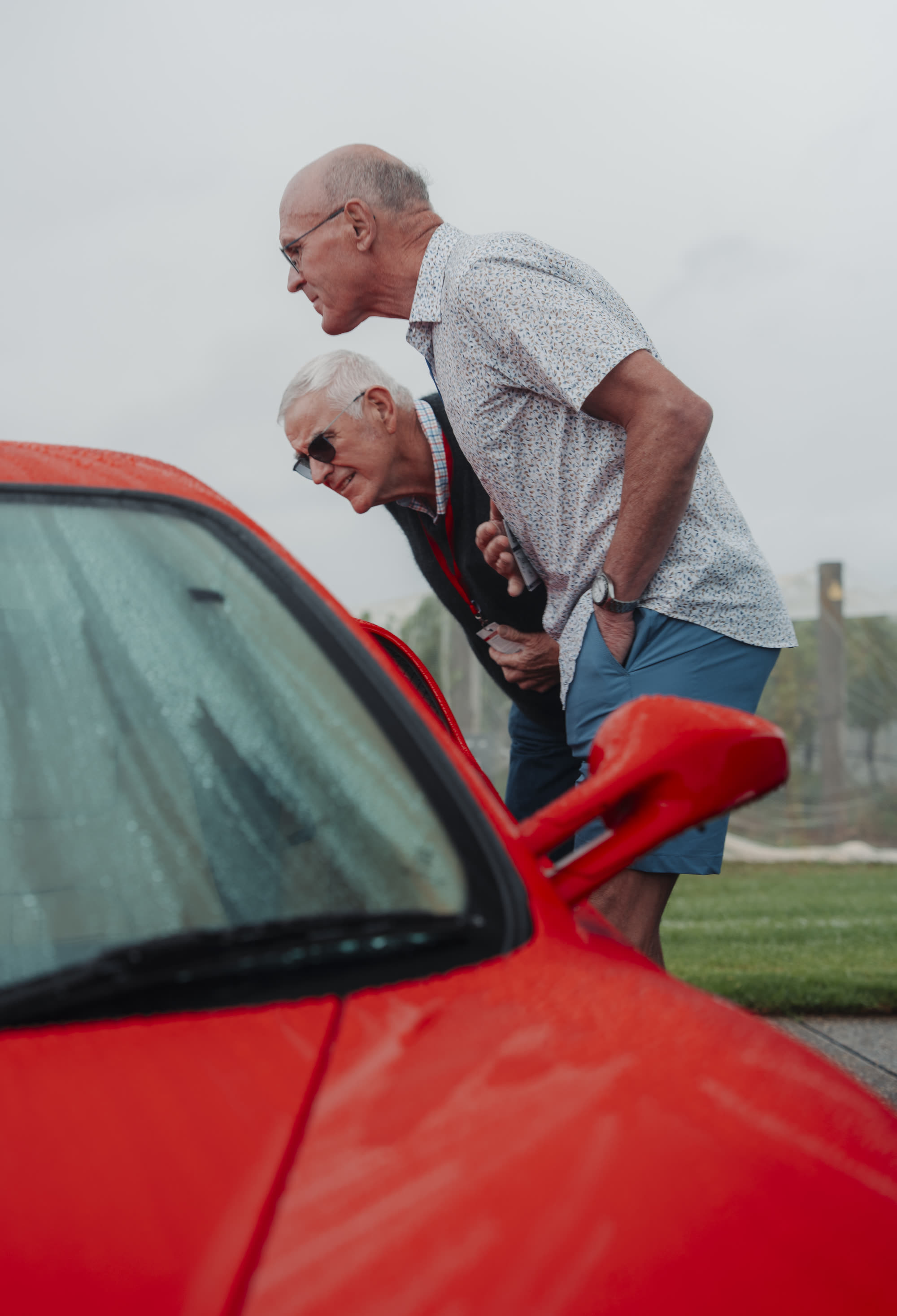 Two older men leaning in to look over a red sports car