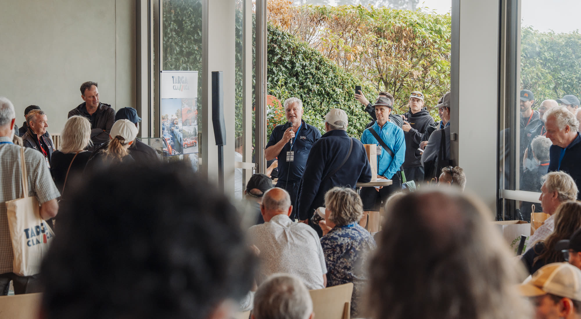 A man with a microphone briefing a seated crowd at a Targa Classica gathering
