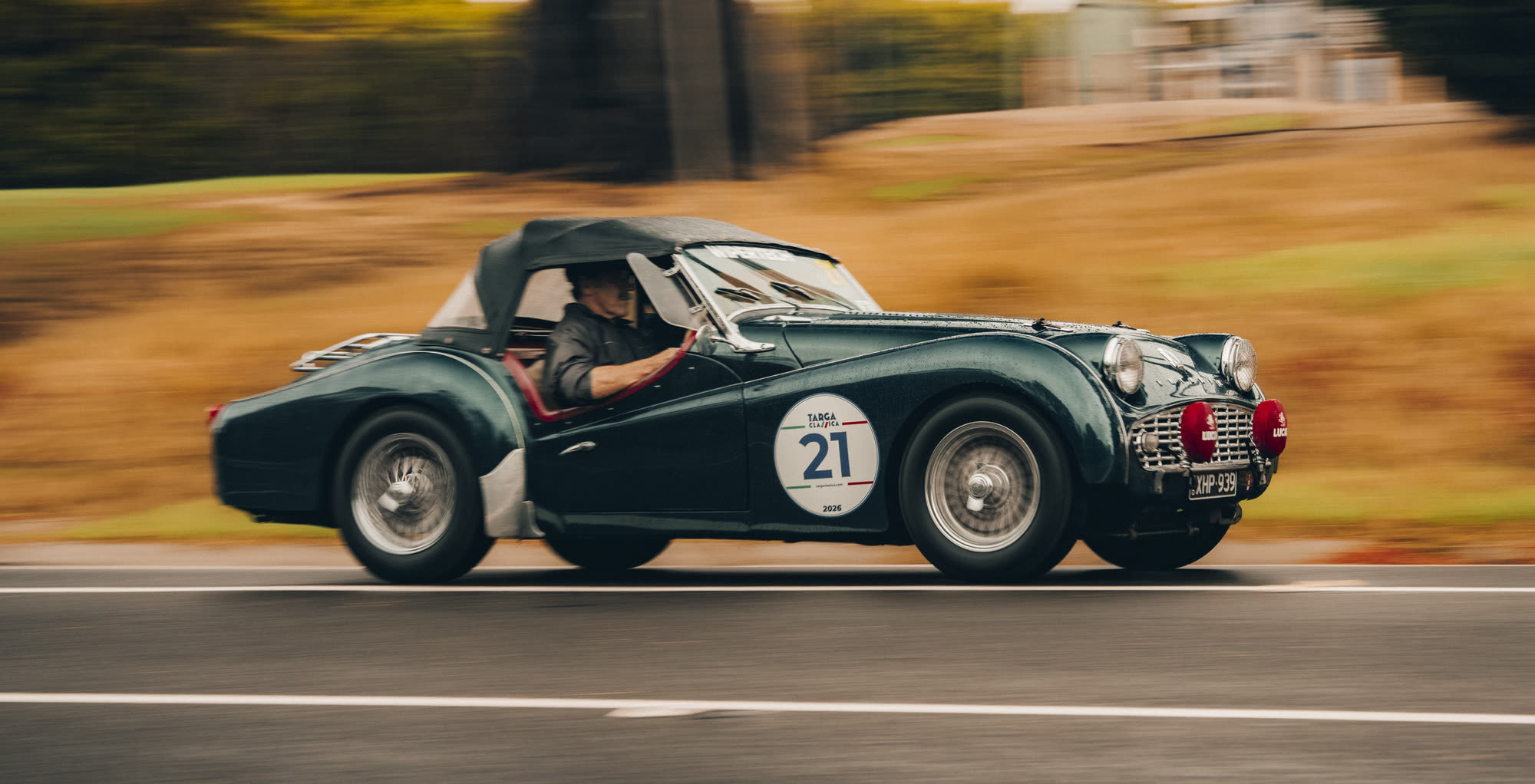 A dark green open-top classic roadster at speed, rally number 21, panned blur
