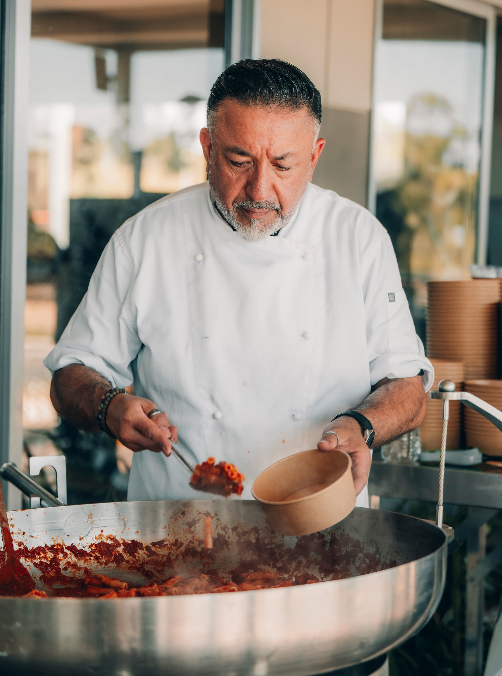 A chef in whites serving pasta from a large pan into a takeaway bowl