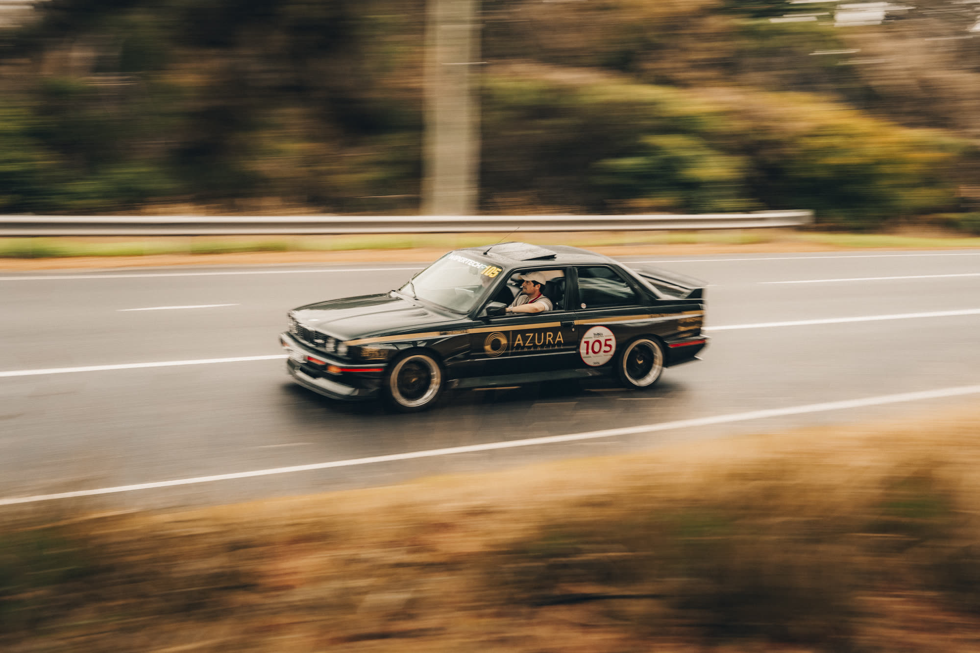 A black BMW coupe with number 105 at speed on a damp country highway