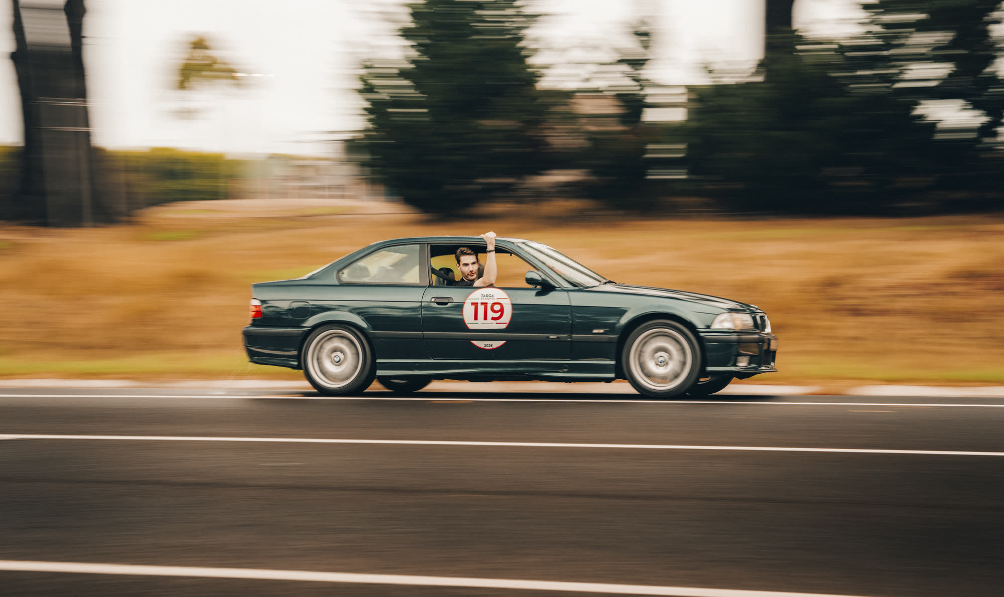 A dark green BMW coupe at speed, driver waving an arm out the window