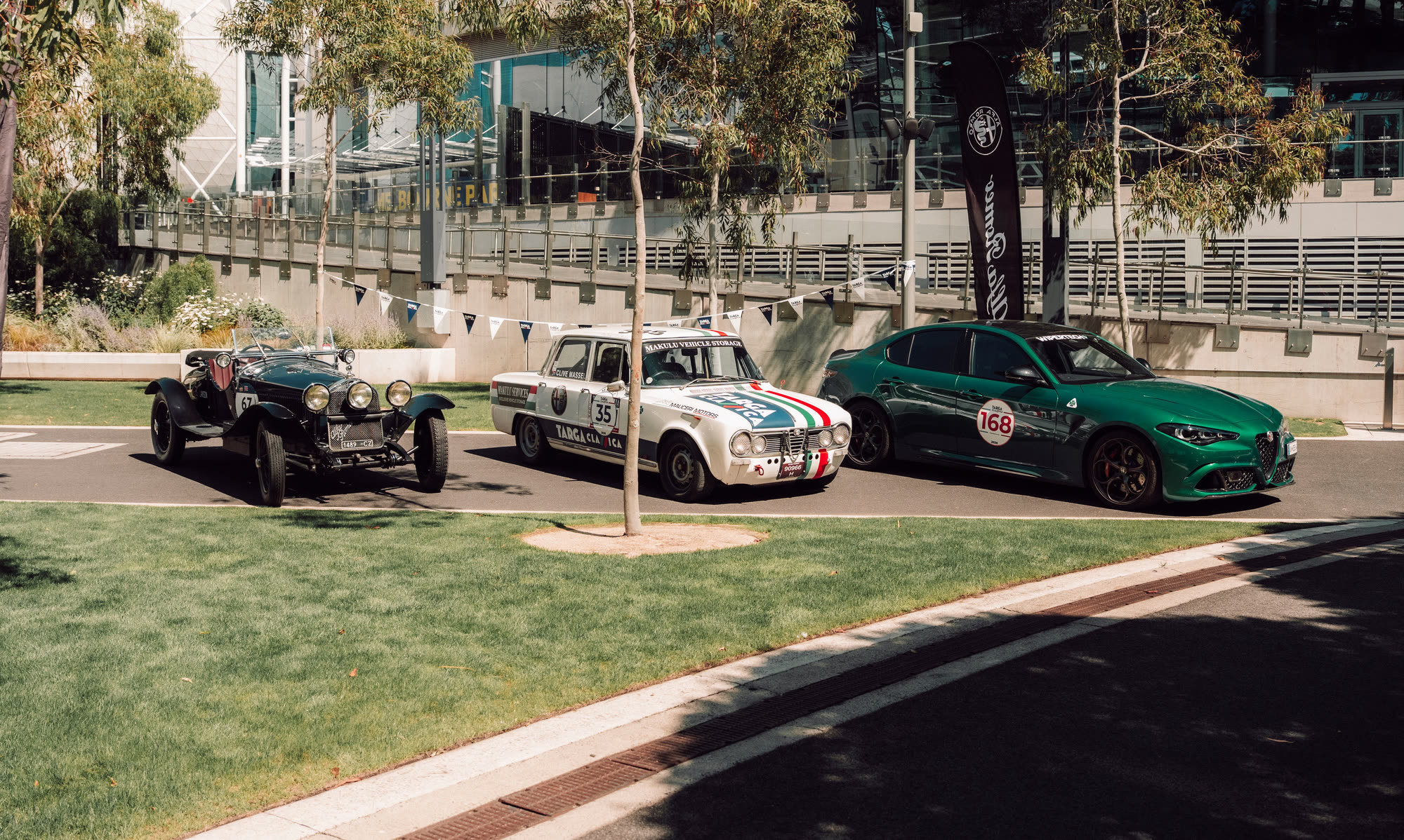 A pre-war tourer, a white rally saloon and a green Alfa Romeo parked kerbside
