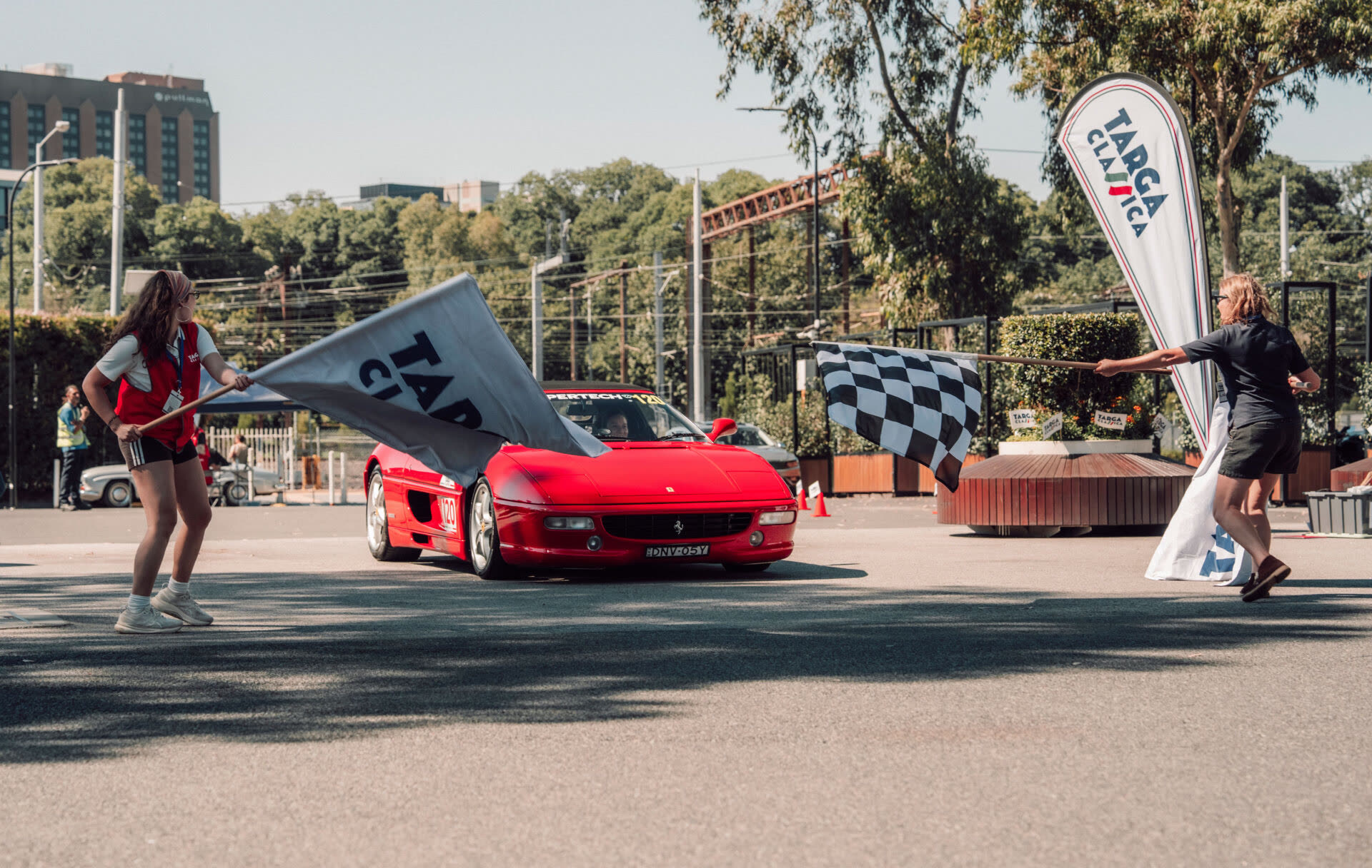 A red Ferrari at the start line as two officials wave Targa Classica flags