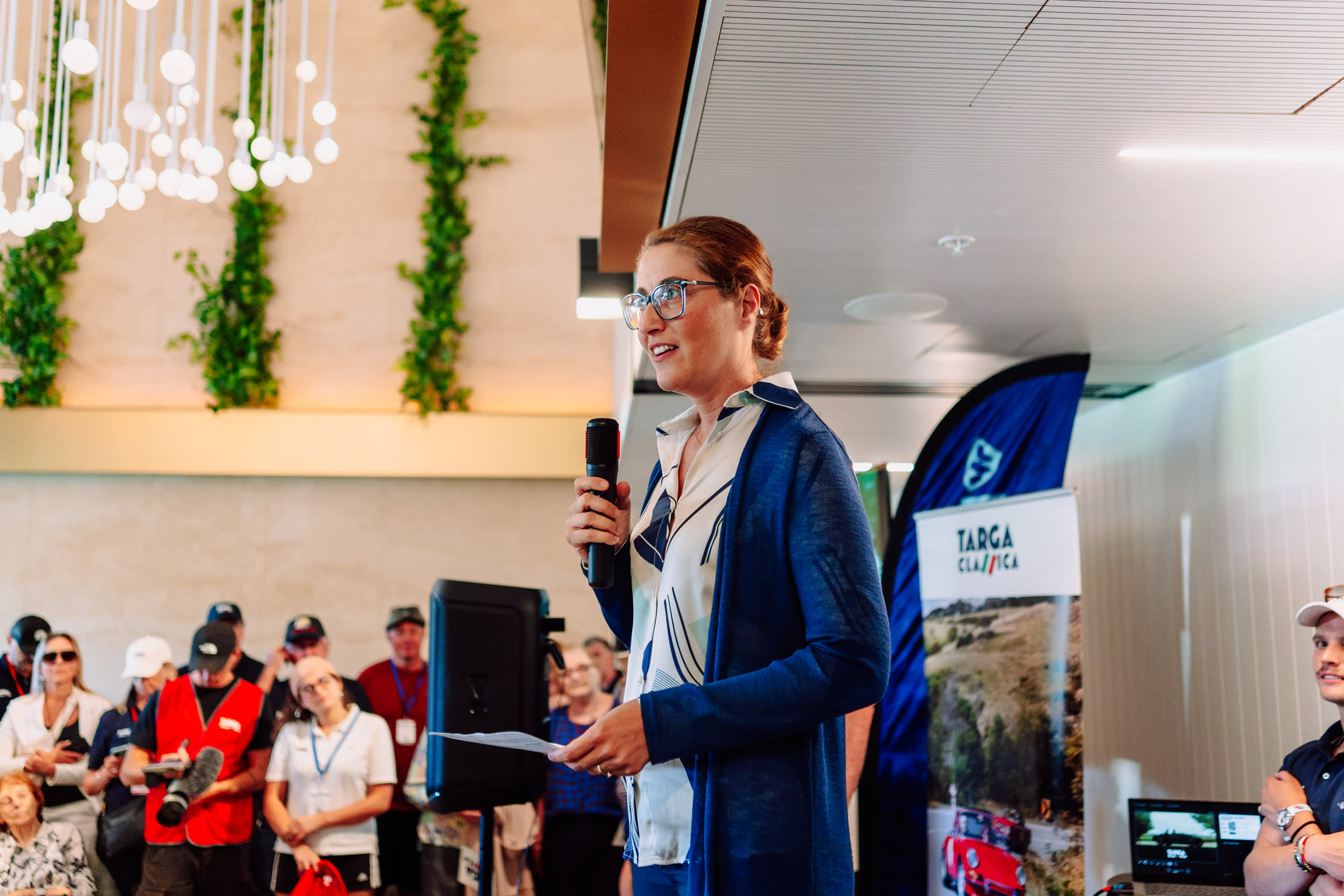 A woman speaking into a microphone to a standing crowd at an event briefing
