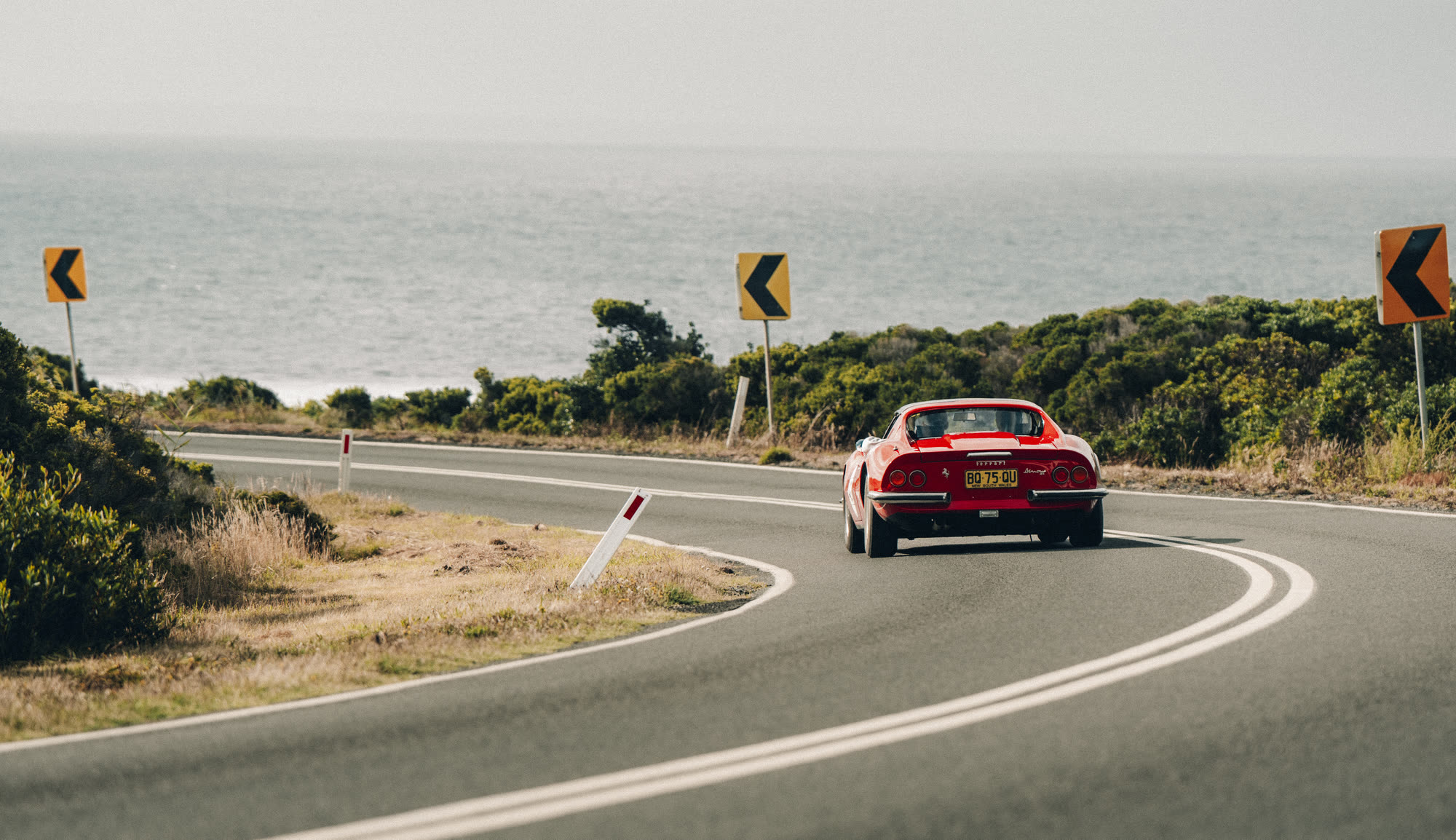 A red Ferrari Dino rounding a bend on a coast road above the sea