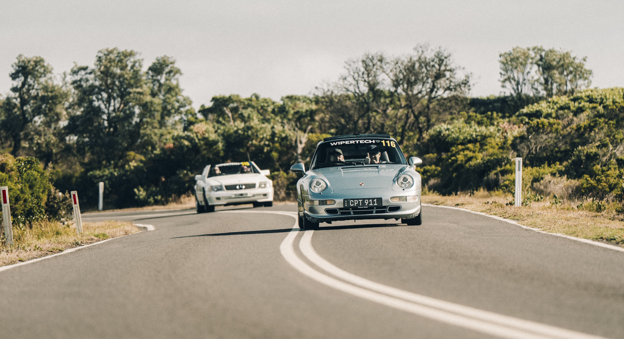 A pale blue Porsche 911 leading a white saloon along a coastal road