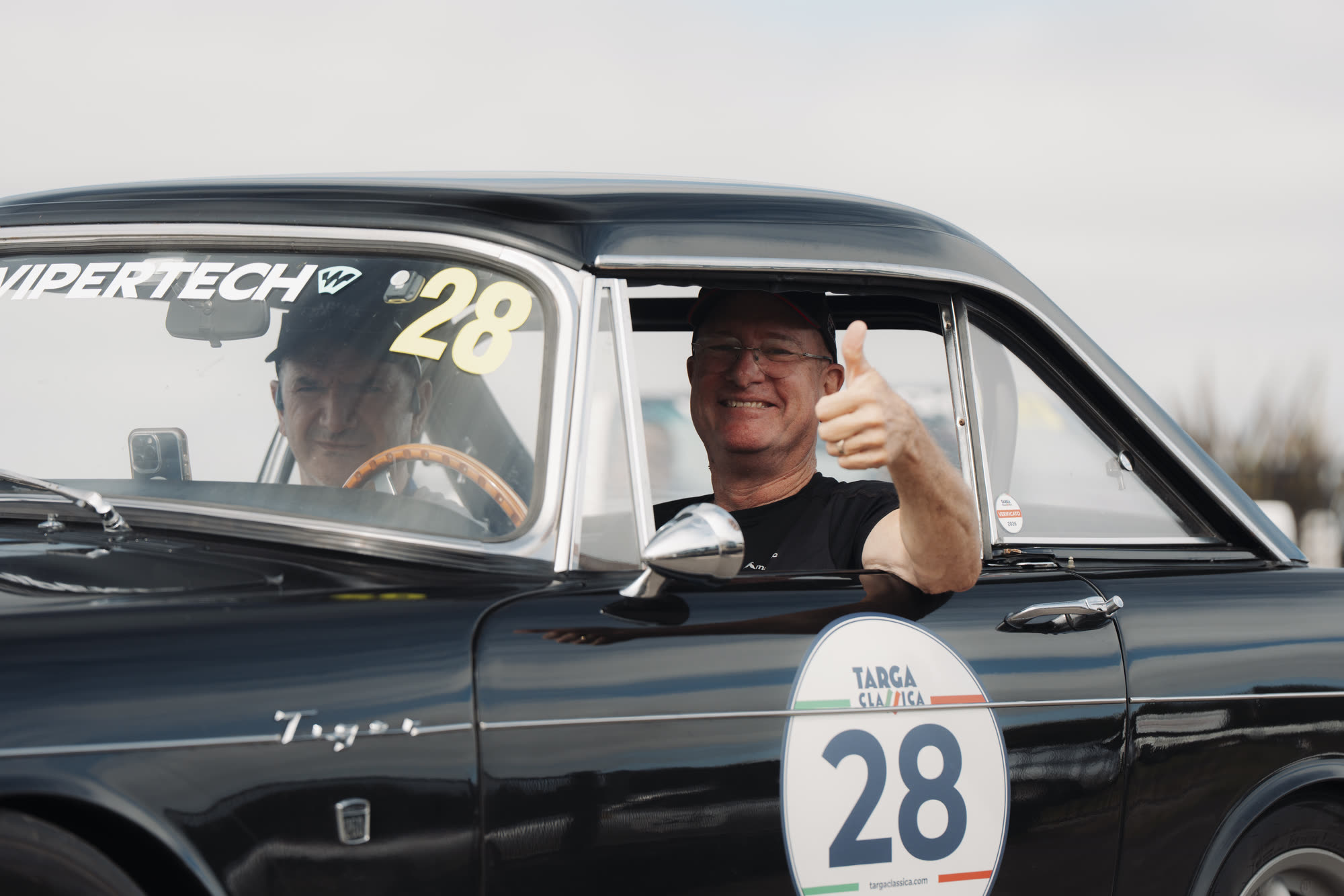Two crew in a black classic saloon, the passenger giving a thumbs up
