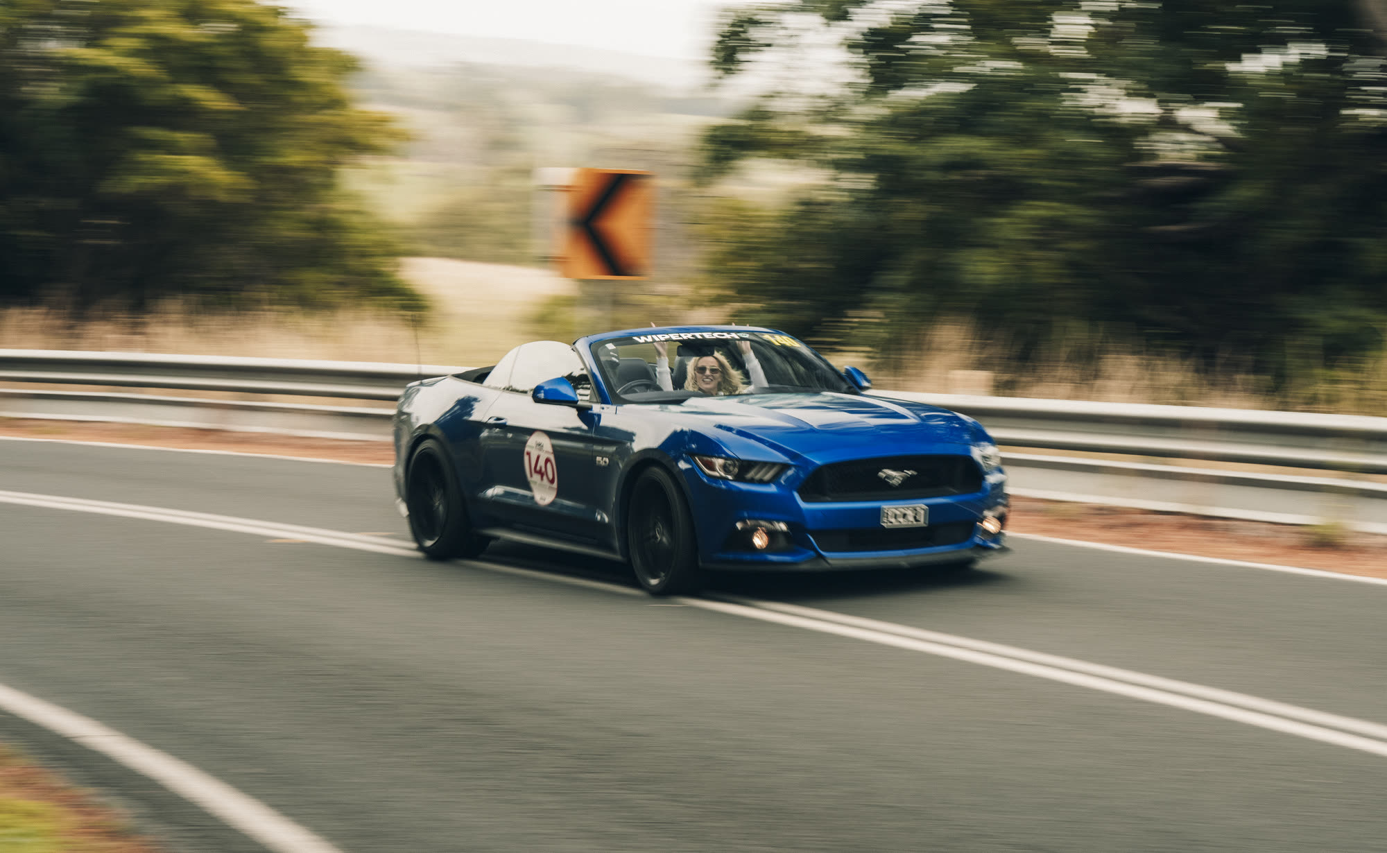A blue Ford Mustang convertible at speed on a country road, number 140