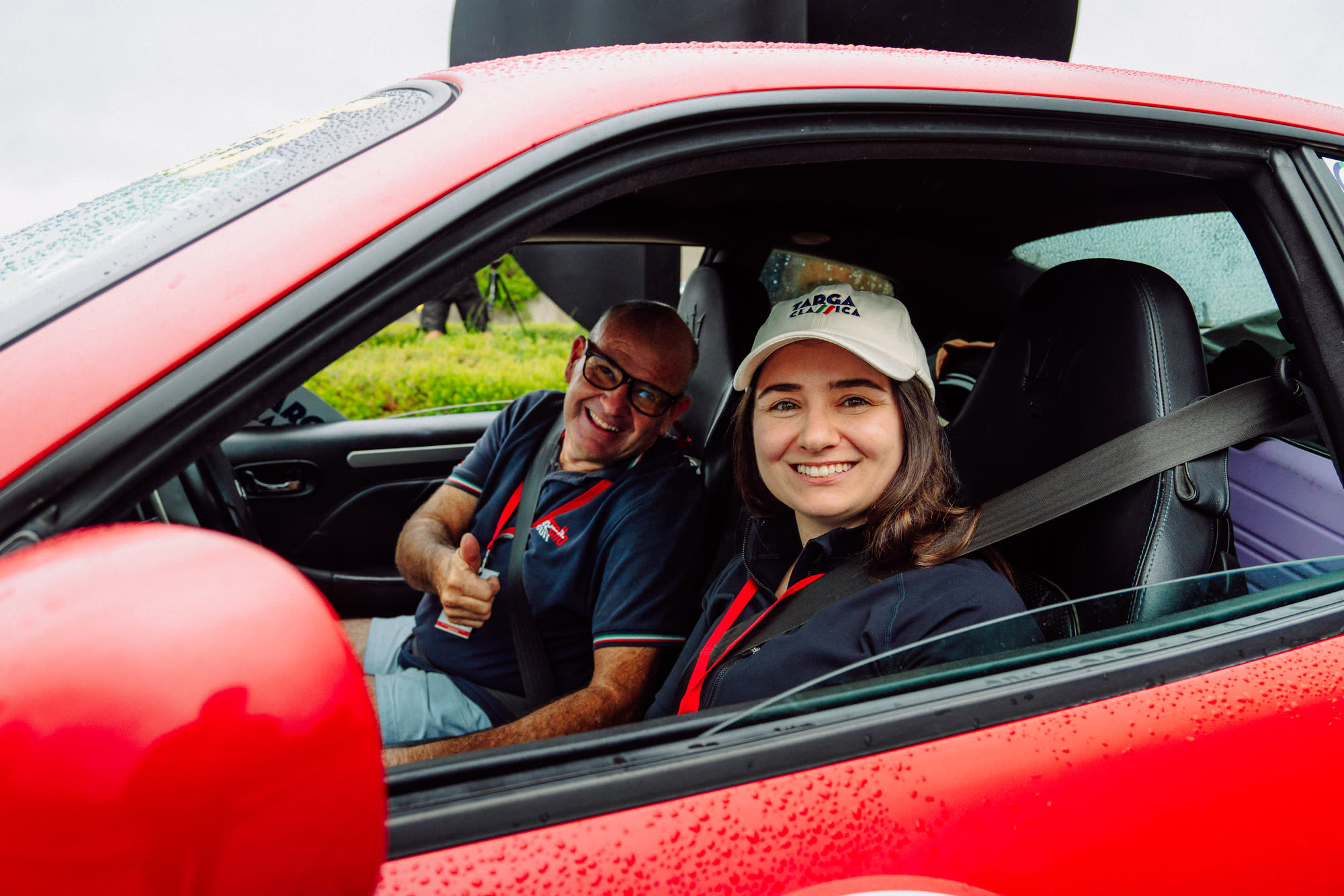 A smiling crew of two in a red car, the driver in a Targa Classica cap