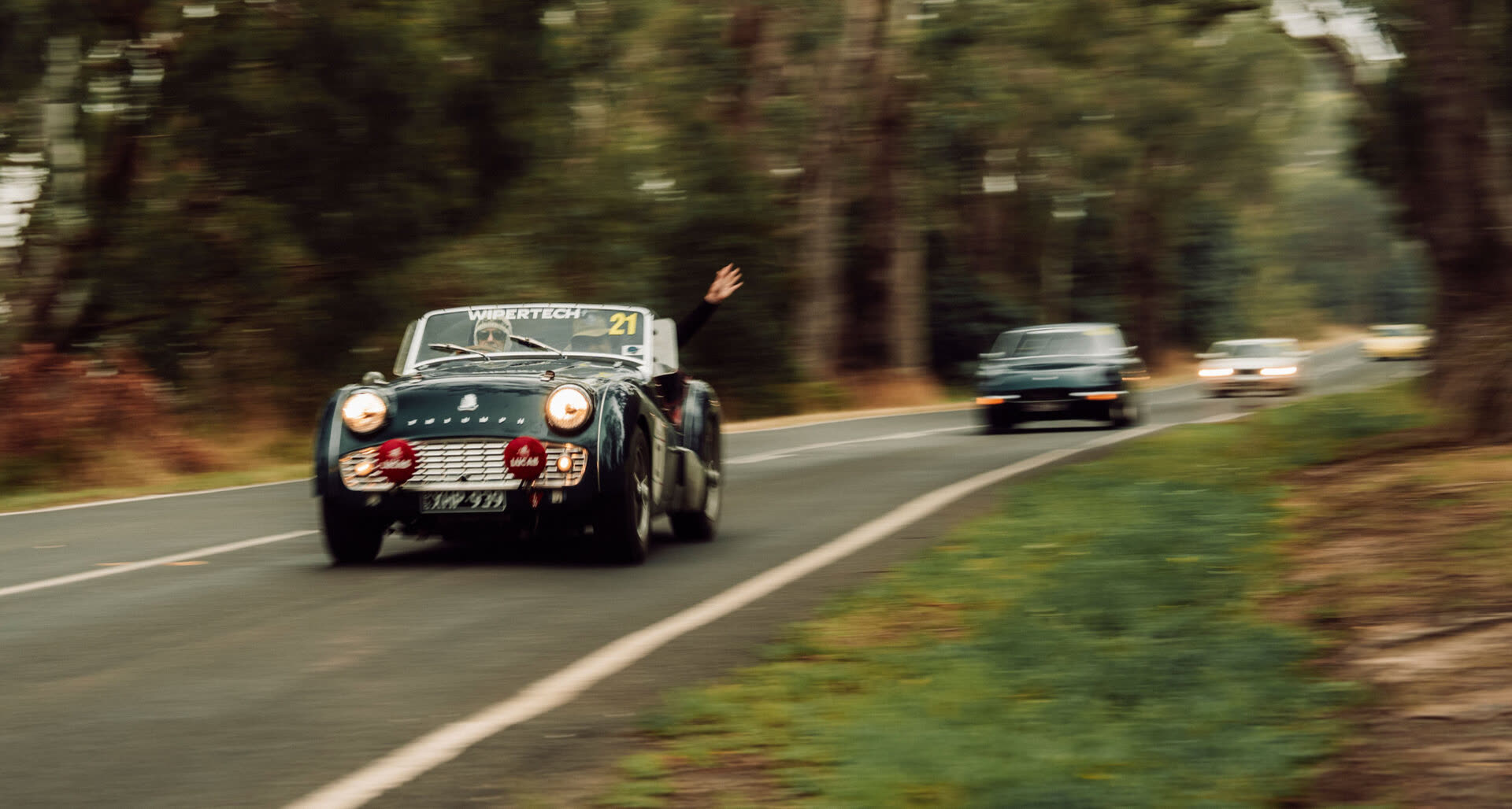 A dark green classic roadster leading a line of cars through forest, arm raised