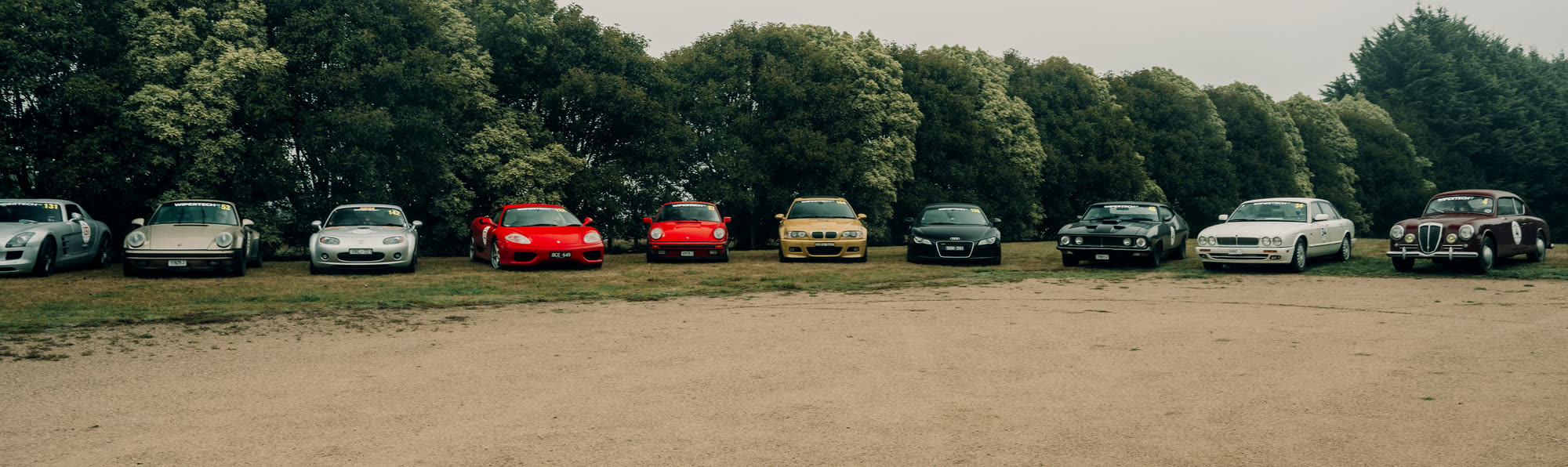 A long line of classic and modern cars parked on gravel before a treeline