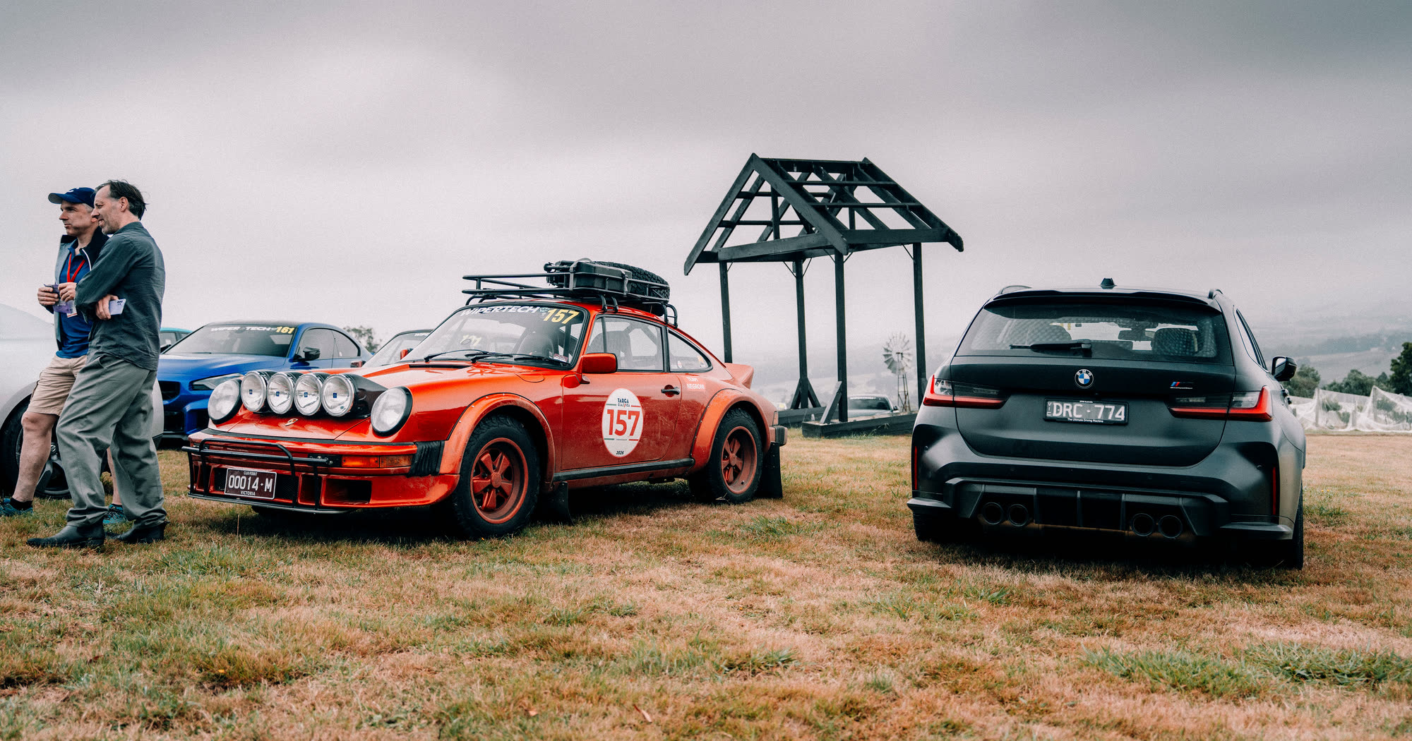 An orange Porsche 911 on a roof rack build parked beside a black BMW wagon